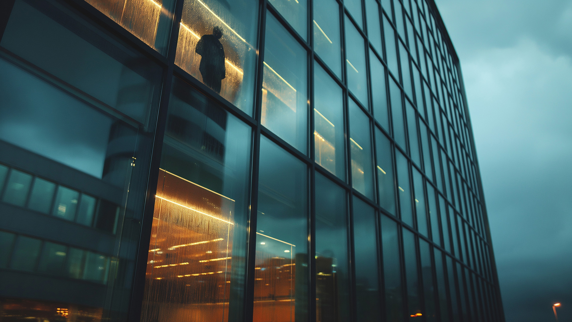 Glass office tower at dusk with one silhouetted figure standing at a lit window