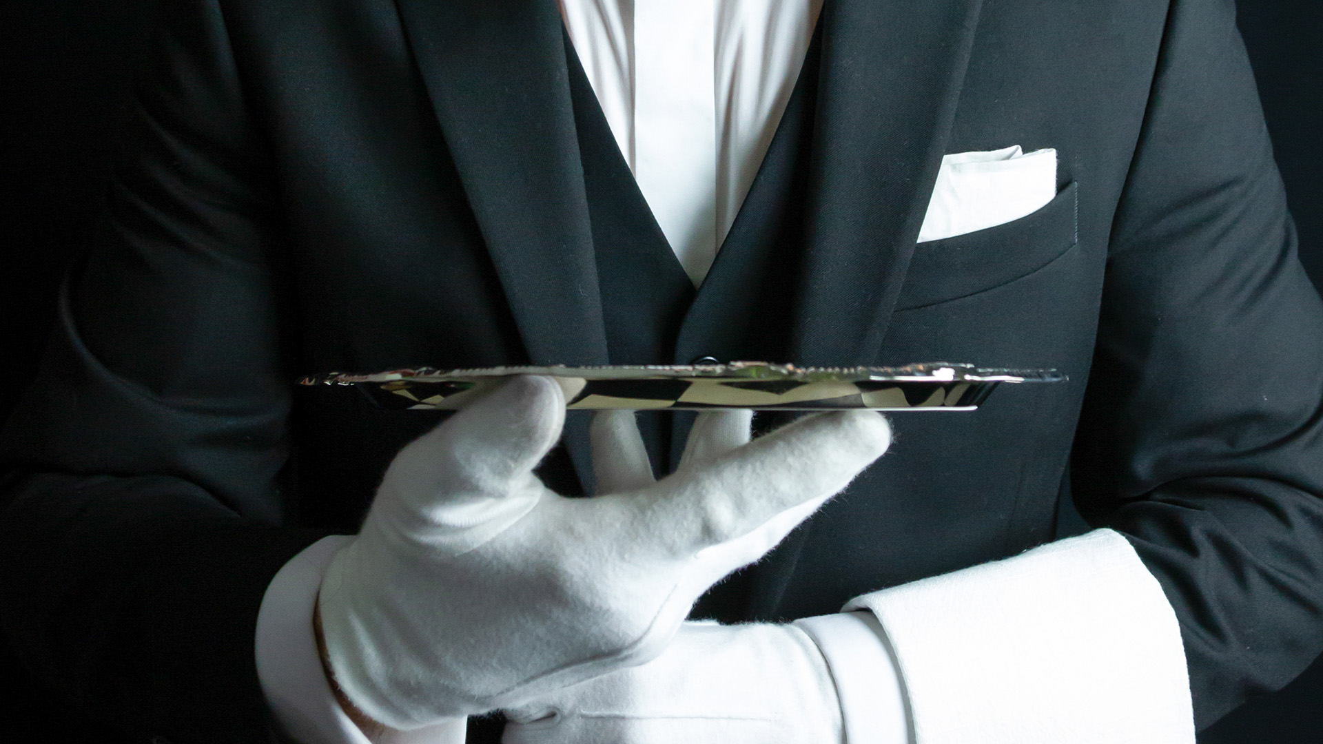 White-gloved hands holding a silver tray in front of a dark tailored jacket